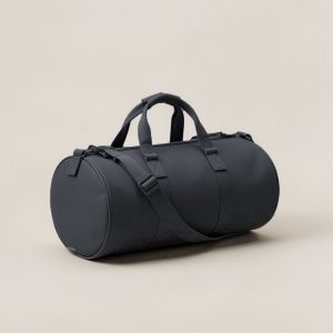Cedar Duffel in Charcoal — cylindrical duffel bag with carry handles and a shoulder strap on a light grey backdrop