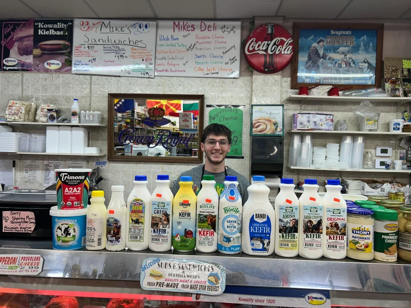 Mike's Deli counter with handwritten sandwich and cold cut boards and a row of kefir and yogurt drinks