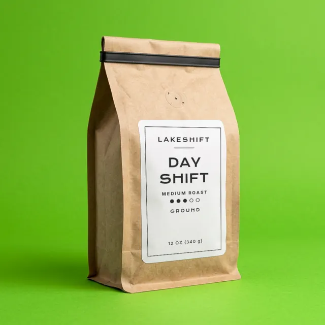 A kraft paper coffee bag standing on a green ground. Its white label reads Lakeshift, Day Shift, medium roast, ground, 12 oz (340 g).