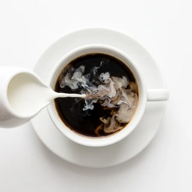 A white cup of black coffee seen from above on a white saucer, with milk being poured in from a small jug.