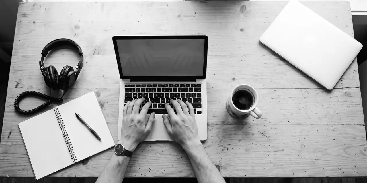 A wooden work desk seen from above: two hands typing on a laptop, an open notebook and pen, headphones and a cup of black coffee.