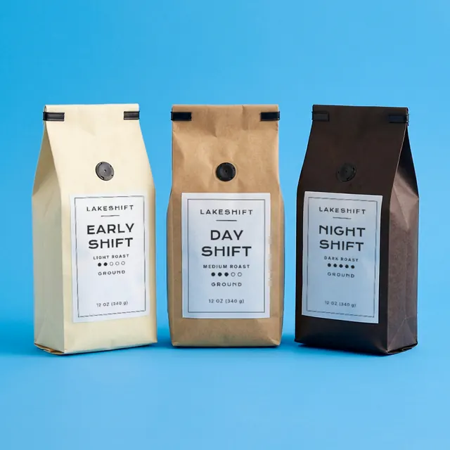 Three Lakeshift coffee bags side by side on a blue ground: Early Shift light roast in cream paper, Day Shift medium roast in kraft paper, Night Shift dark roast in dark brown paper.